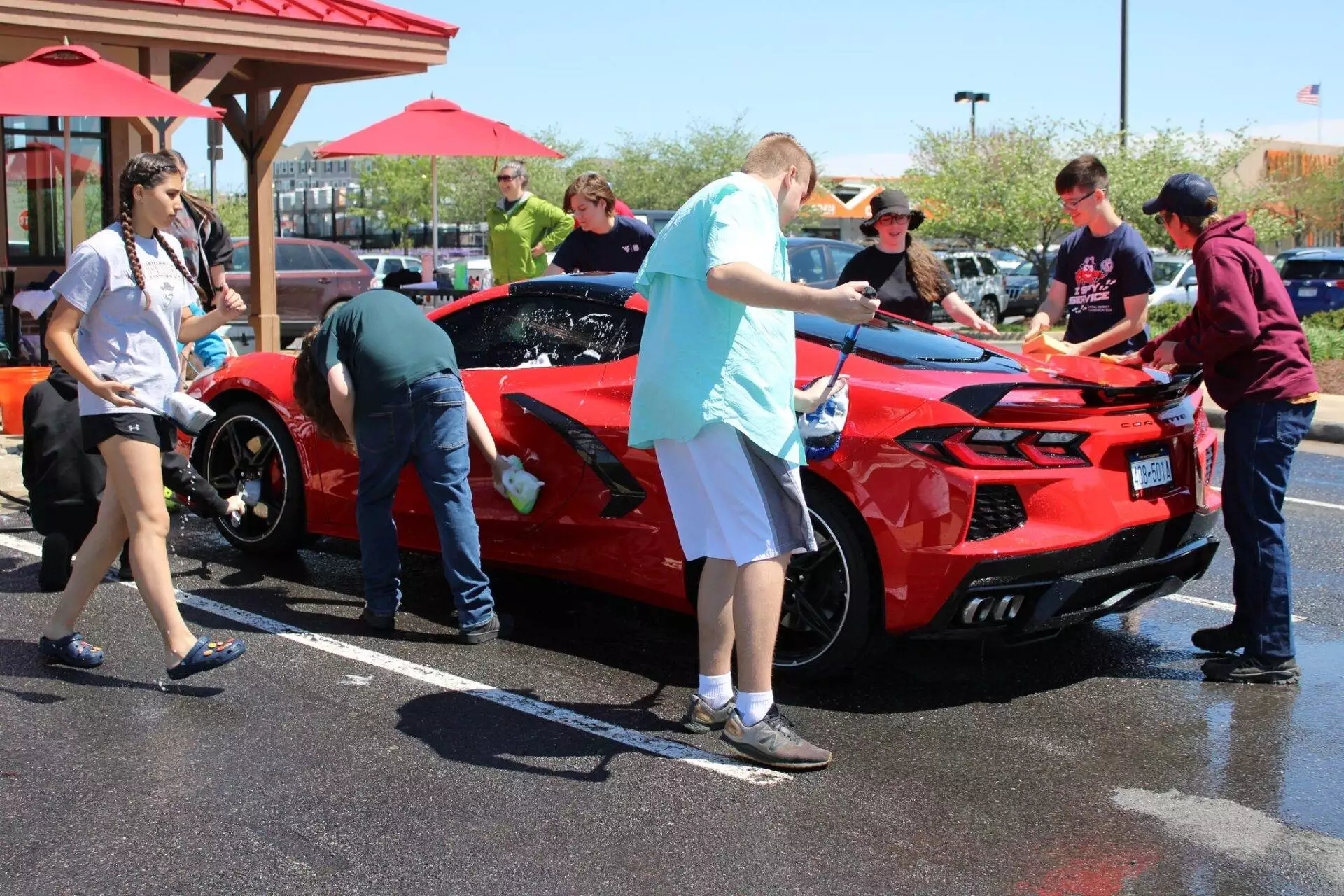People washing a red sports car in a sunny parking lot, with one person wiping the hood and others cleaning various parts of the car.