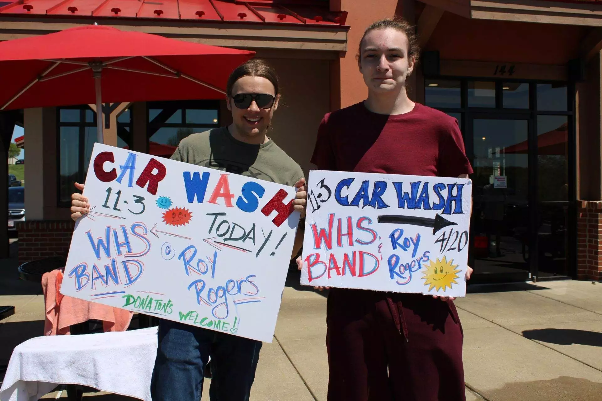 Two people holding handmade signs promoting a car wash fundraiser for a school band, standing in front of a restaurant.