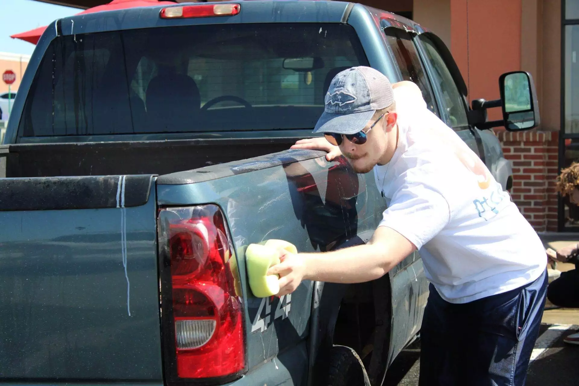 A man in a white t-shirt and cap diligently washing a red pickup truck in a sunny outdoor setting.