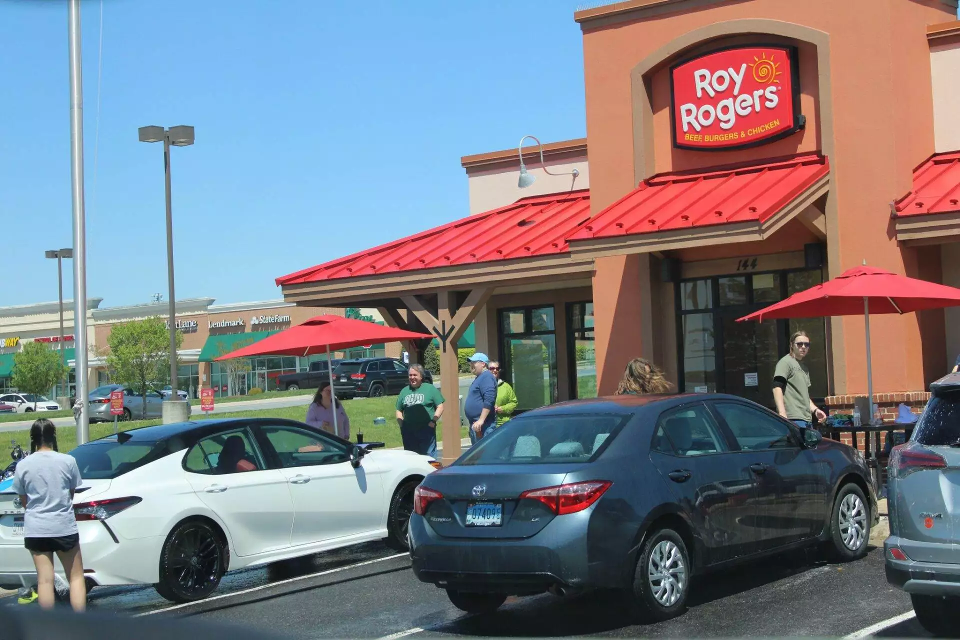 Cars parked outside a roy rogers restaurant on a sunny day, with people walking near the entrance under red umbrellas.