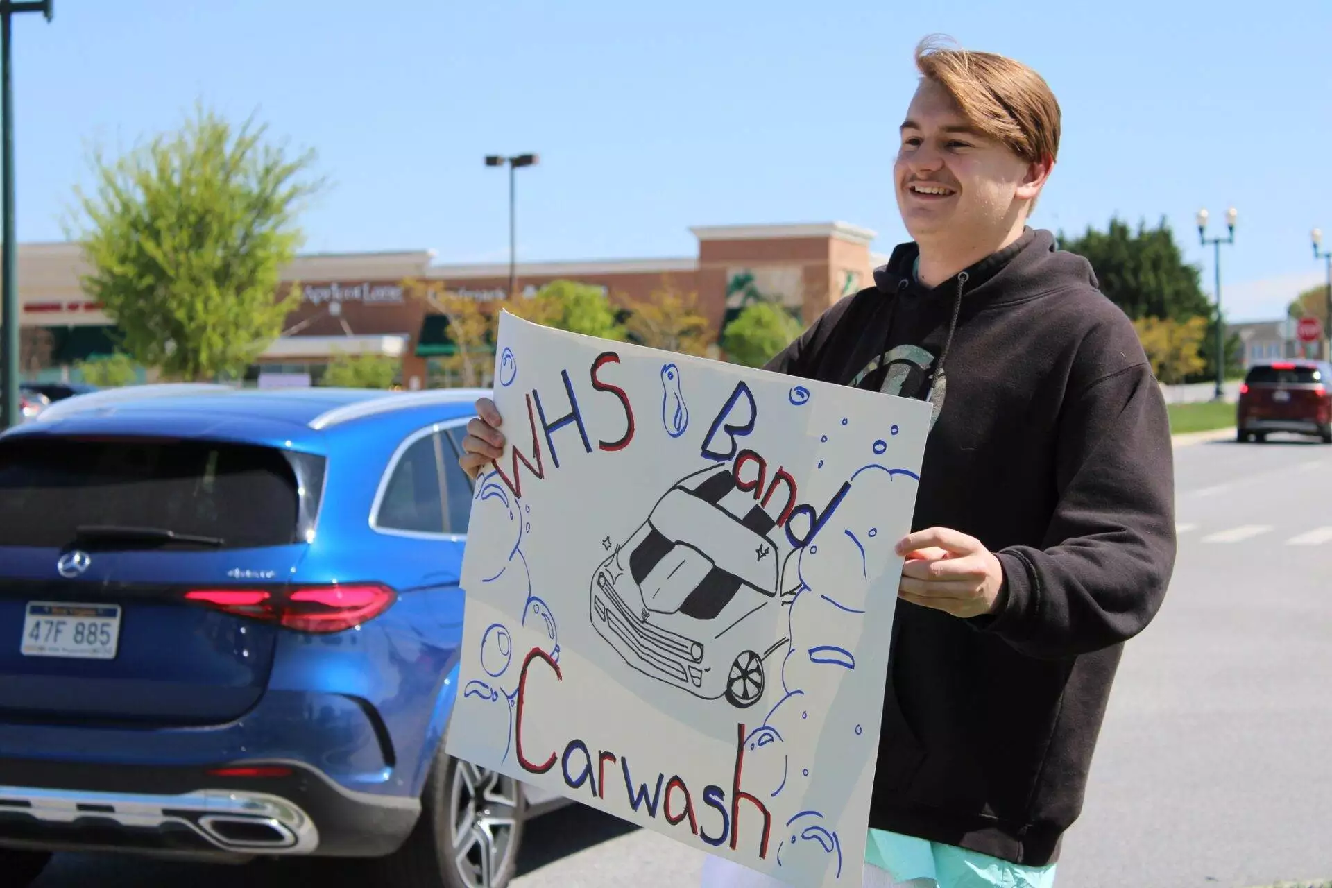 A young man holding a sign advertising a high school band car wash on a sunny day, with cars and a shopping complex in the background.