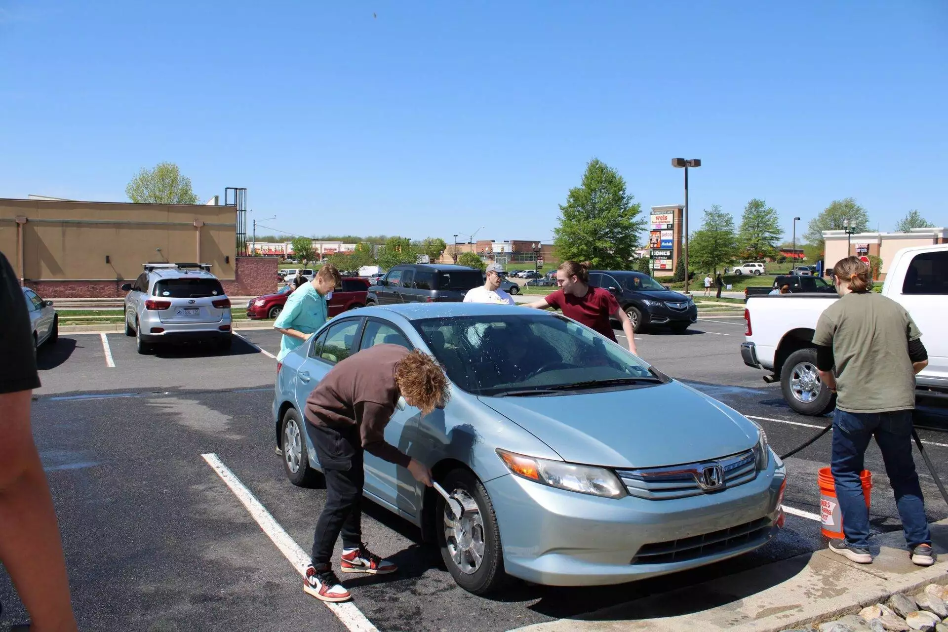A group of people washing cars in a sunny parking lot, with various vehicles and a commercial building in the background.