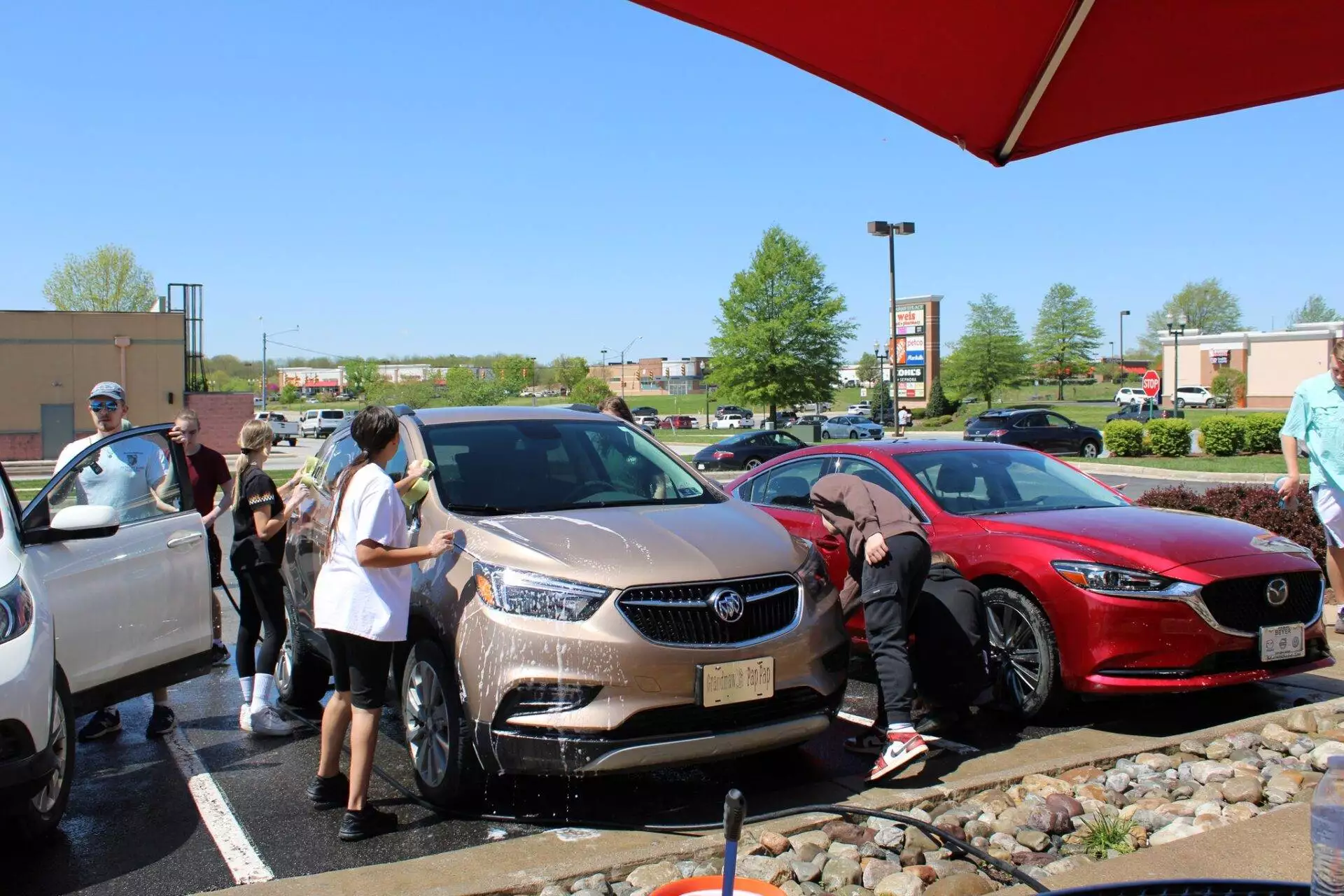A group of people washing cars in a sunny parking lot, including a tan sedan and a red sports car.