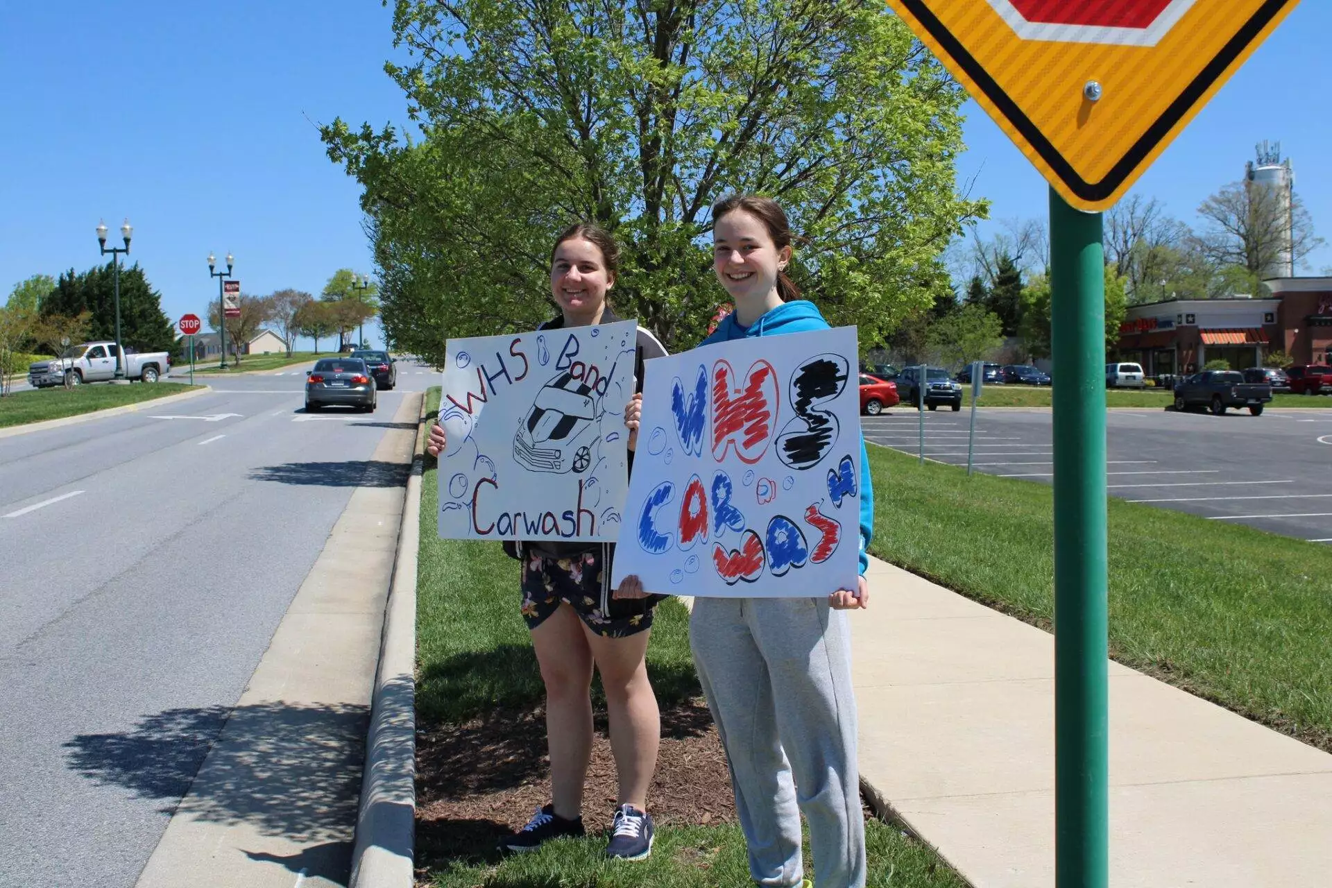 Two teenagers holding signs advertising a car wash fundraiser by the roadside on a sunny day.