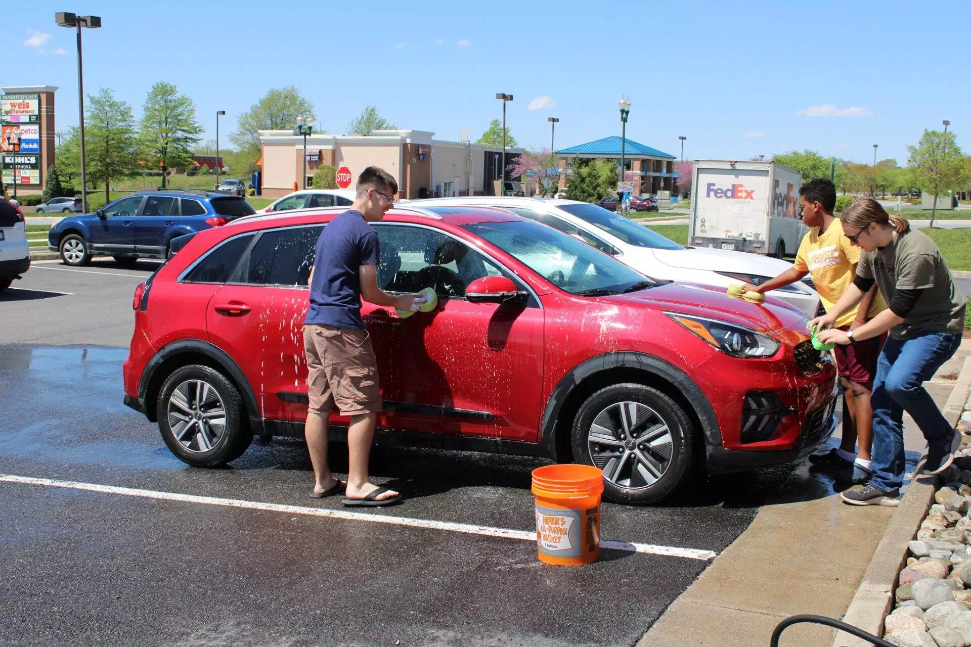 Three people washing a red suv in a sunny parking lot, using buckets and sponges.
