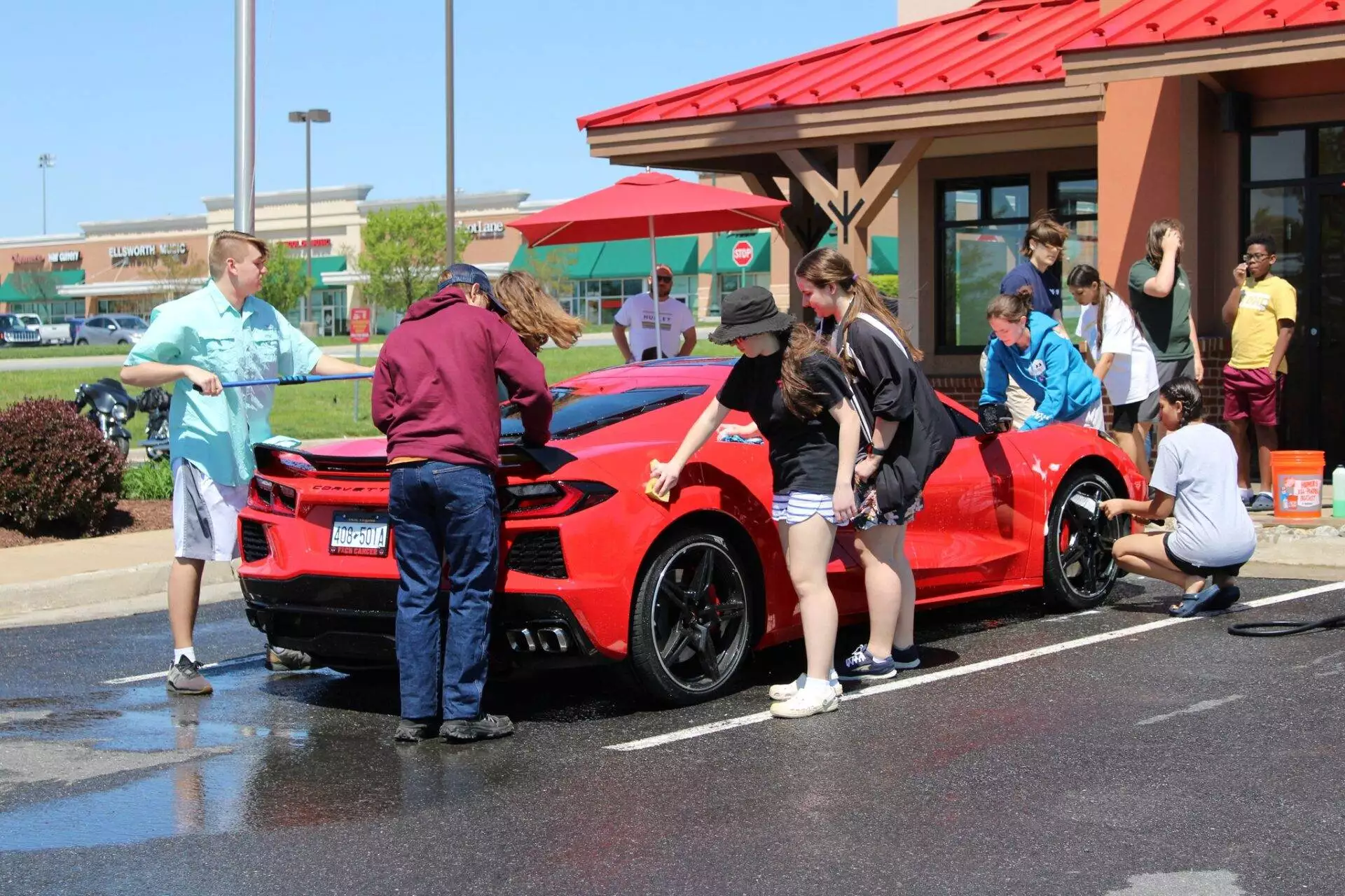 Group of people washing a red sports car in a sunny parking lot outside a building.